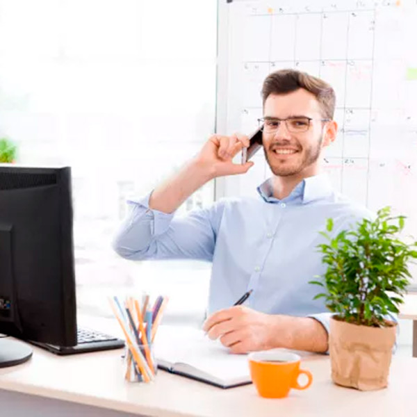 Chico joven con gafas habla por el móvil en su escritorio con ordenador de sobremesa
