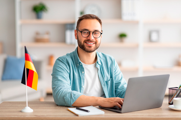 Chico delante de un portátil en un escritorio con una bandera de alemania, aprende alemán en un curso online.