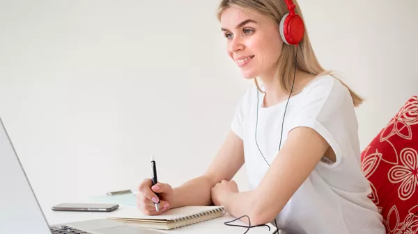 Chica rubia con auriculares mirando hacia un monitor y tomando notas en una libreta
