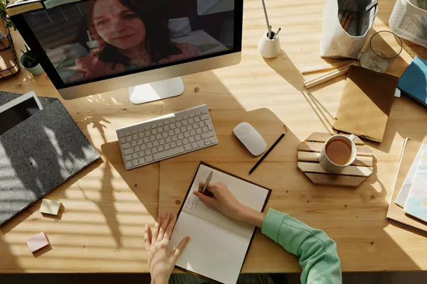 Escritorio con una película en un monitor, un café y unas manos de mujer tomando notas en una libreta