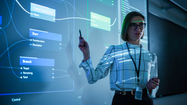 Profesora de universidad, rubia con gafas y camisa blanca de cuadros grandes en negro da una clase sobre big data delante de un proyector que refleja en la pizarra varios diagramas con flujos de información