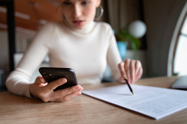 Repasa los datos de contacto al escribir tu carta de presentación chica joven desenfocada, tiene el móvil en una mano y un folio con bolígrafo