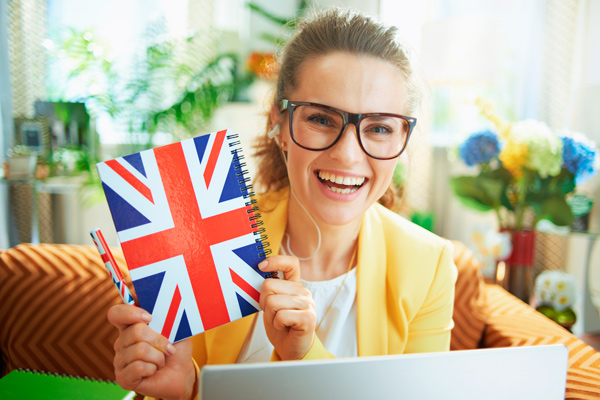 Cómo estudiar inglés B2 con Femxa chica joven, rubia con gafas con una libreta con la bandera del Reino Unido, estudia inglés en un portátil.