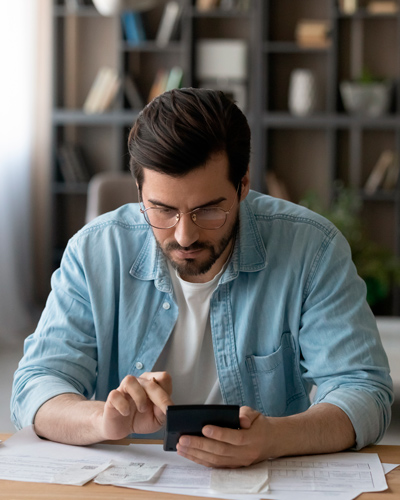 chico con gafas, camisa azul abierta y camiseta blanca con una calculadora en las manos y papeles, está calculando el importe de sus nóminas para tratar de entenderlas