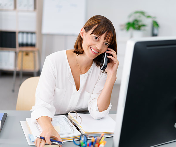 Chica con flequillo y sonrisa con camisa blanca atiende el teléfono con un fichero de papeles y un ordenador de sobremesa