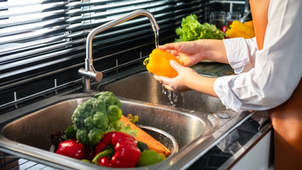 manos de mujer lavando verduras en un fregadero de acero inoxidable para hacer recetas que eviten la deshidratación