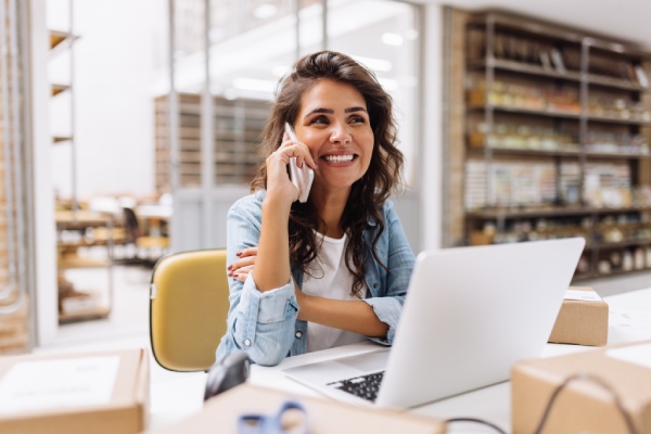 trabajadora autónoma gestionando su empresa por teléfono desde un escritorio, al fondo se ven estanterías con productos y cajas