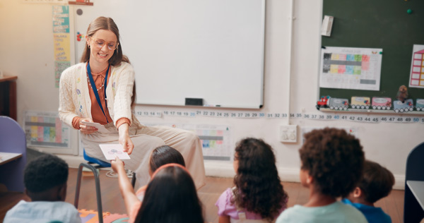 Profesora de pelo largo y gafas redondas con la pizarra al fondo, entrega tarjetas a los niños en un aula