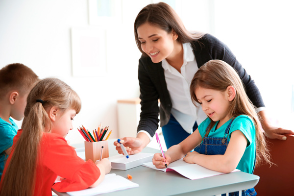 Proceso de aprendizaje de lectoescritura Profesora en el proceso de aprendizaje de lectoescritura en una mesa con dos niñas y un niño con lápices de colores escribiendo en una libreta