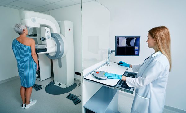 Realización de mamografía, se ve una paciente de espaldas frente a la máquina de radiología y a una técnico sanitaria tras un cristal a los mandos del aparato de mamografía
