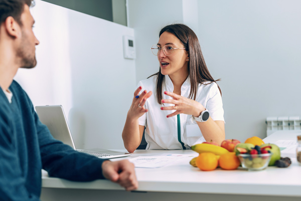 Nutrióloga atendiendo a un paciente, con bata blanca, detrás de una mesa con un portátil y con fruta encima de la mesa
