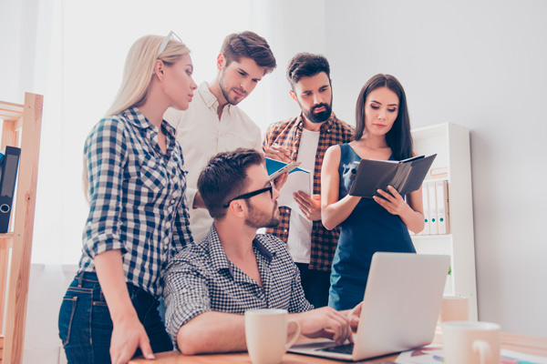 grupo de personas en una empresa aplican el pensamiento crítico, unos de pie tomando notas y otro sentado con el portátil