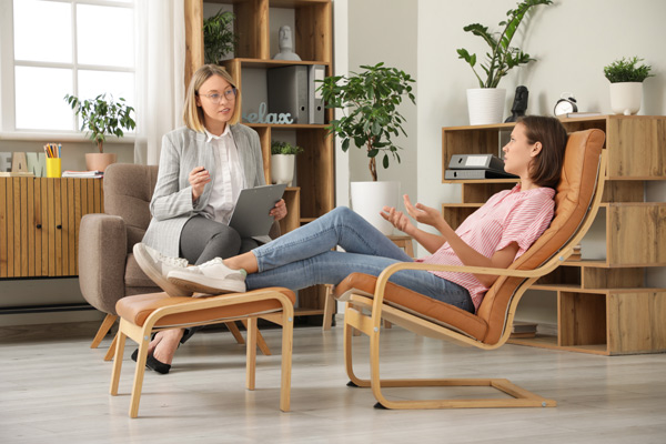Psicóloga joven de pelo rubio y gafas redondas sentada en una butaca atiende a una joven adolescente tumbada en una silla ergonómica