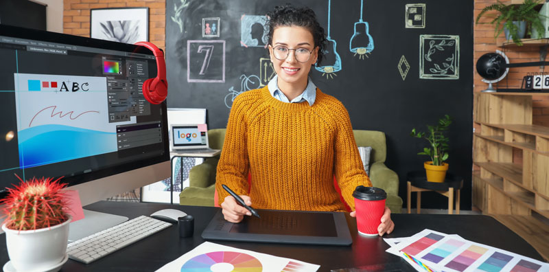 diseñadora editorial, con gafas redondas y jersey color mostaza, en su escritorio con una tarjeta gráfica y un monitor en el que diseña un libro con quarkxpress