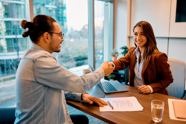 técnica de recursos humanos, joven con pelo largo en su mesa de despacho estrecha la mano de un chico de gafas con moño que acaba de firmar un contrato de trabajo